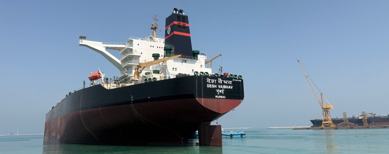Large tanker vessel docked in a harbor, with shipyard cranes in the background.