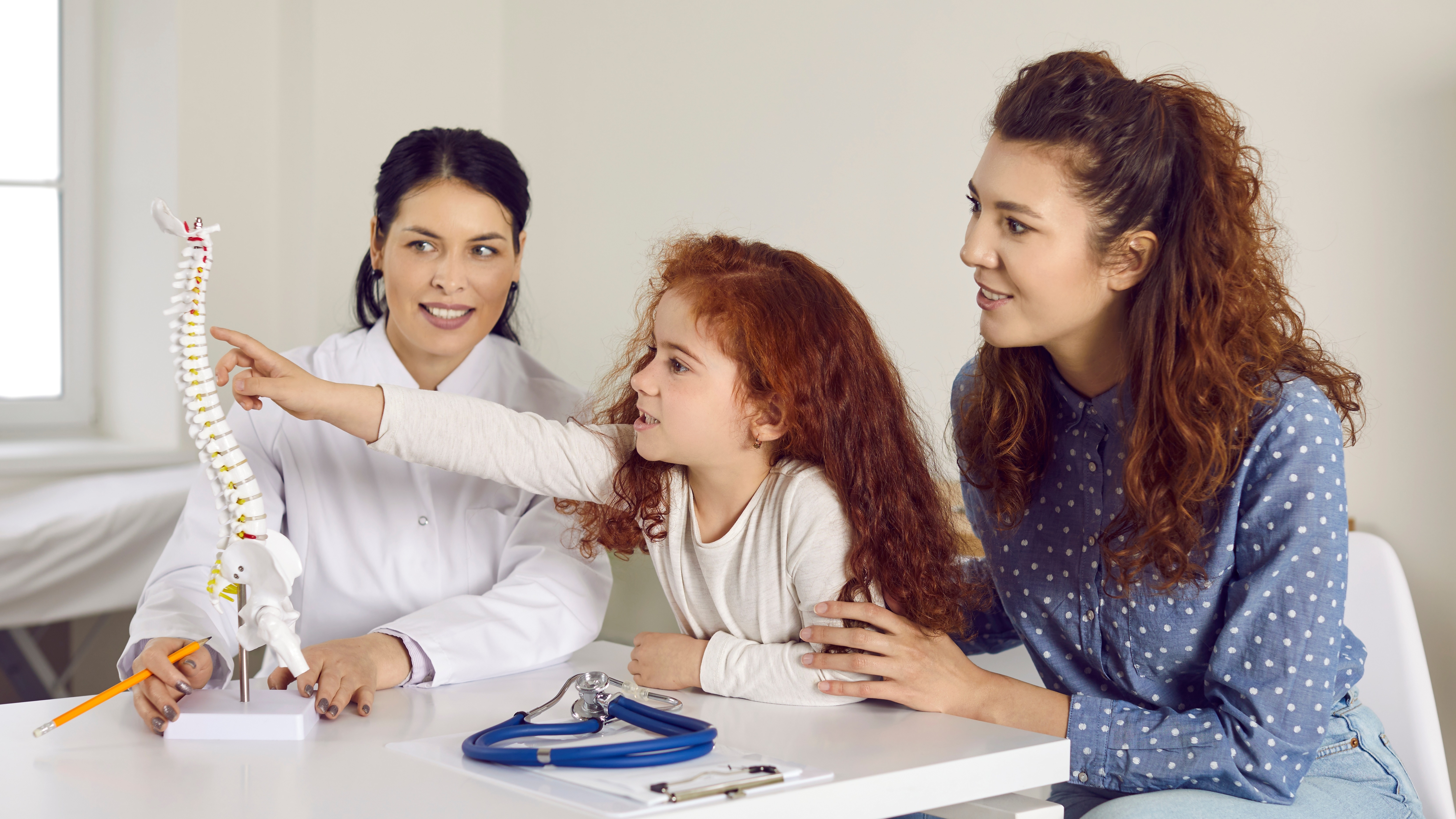 A smiling clinician and parent explain a spine model to a young patient, symbolizing trust, collaboration, and excellence in paediatric spinal care.