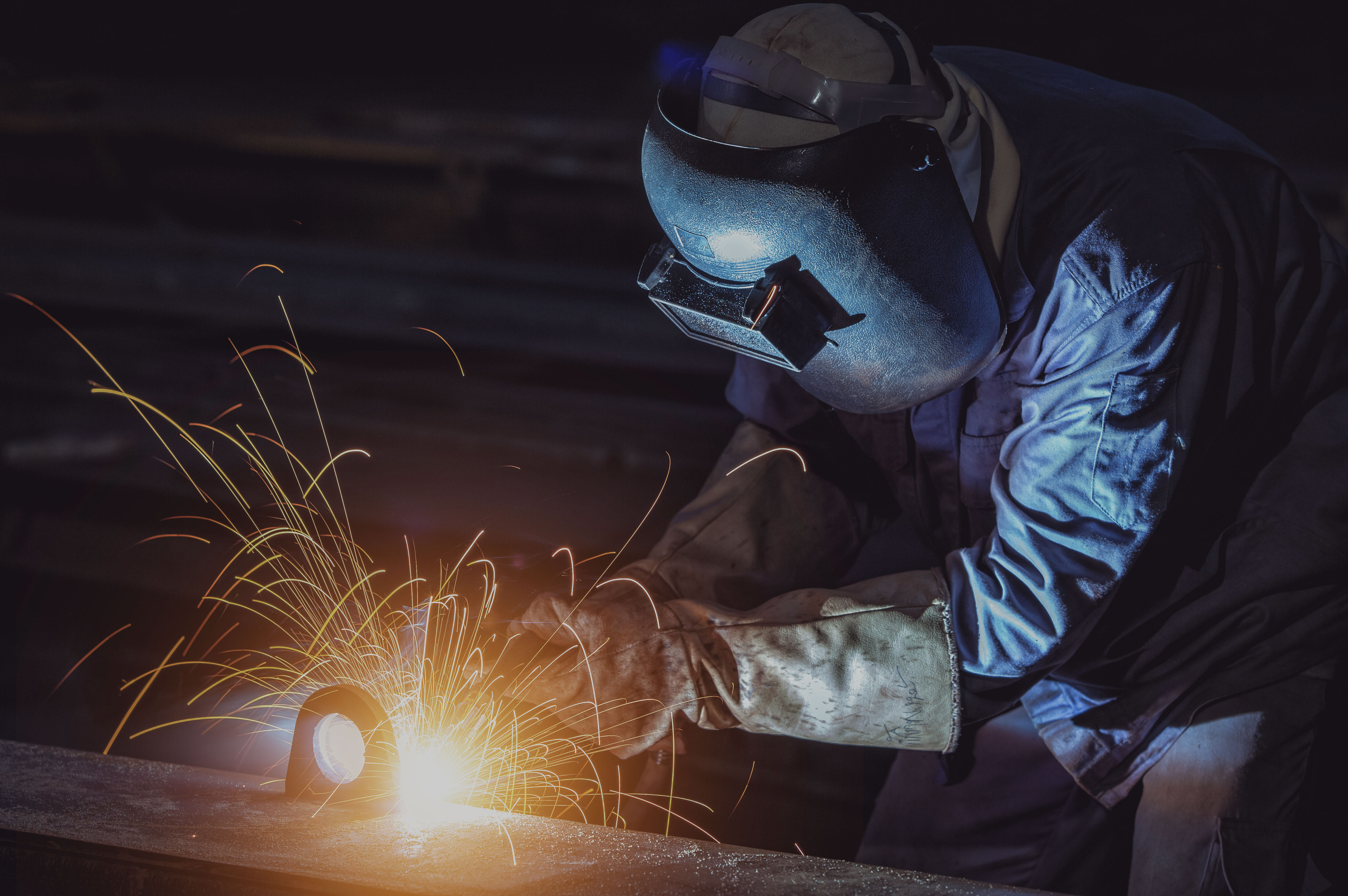 Welder wearing protective gear joining metal components with bright sparks and smoke, illustrating permanent joining and welding operations.