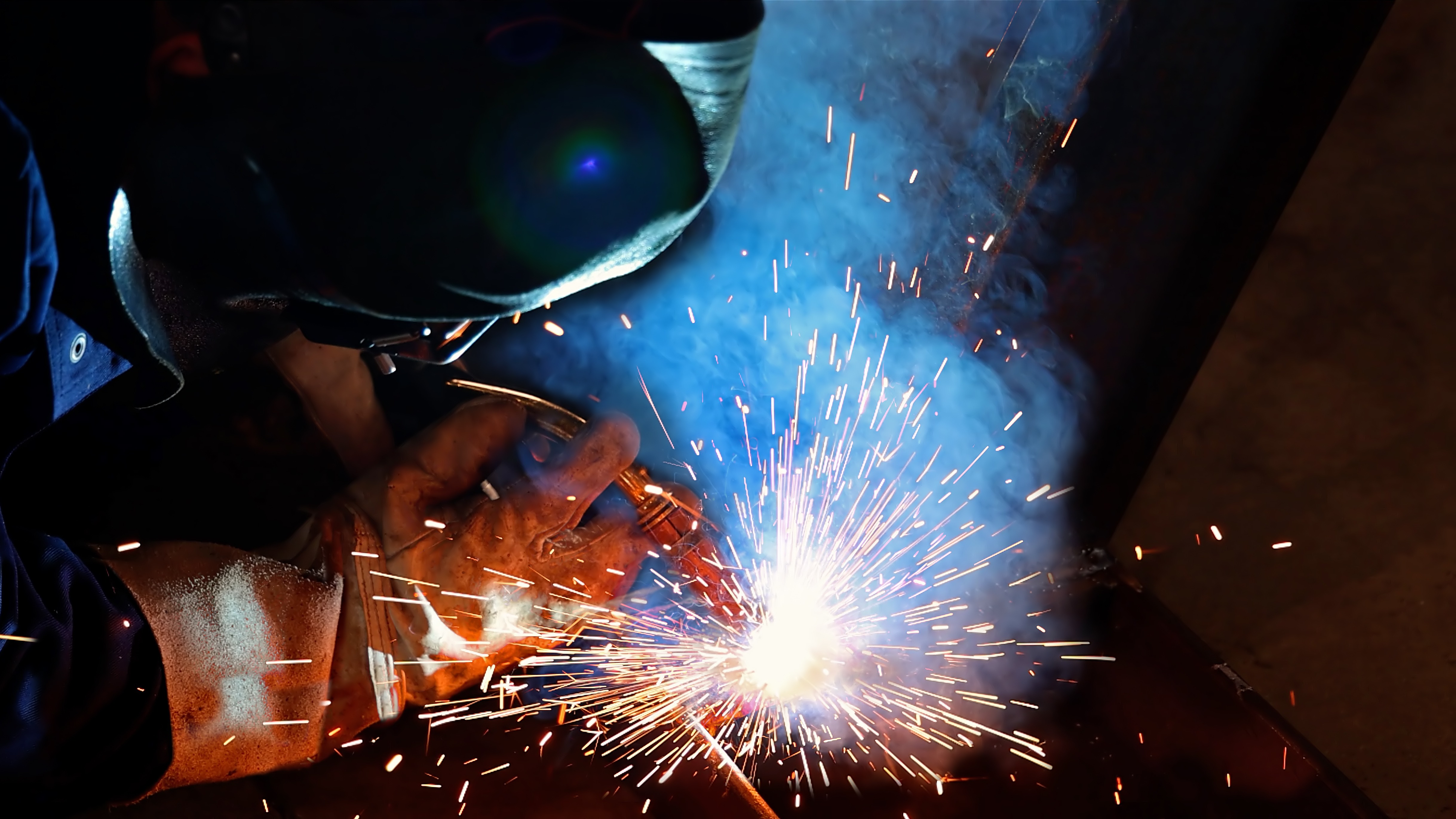 Welder wearing protective gear creating bright sparks while working on metal, illustrating qualified welding operations in an industrial setting